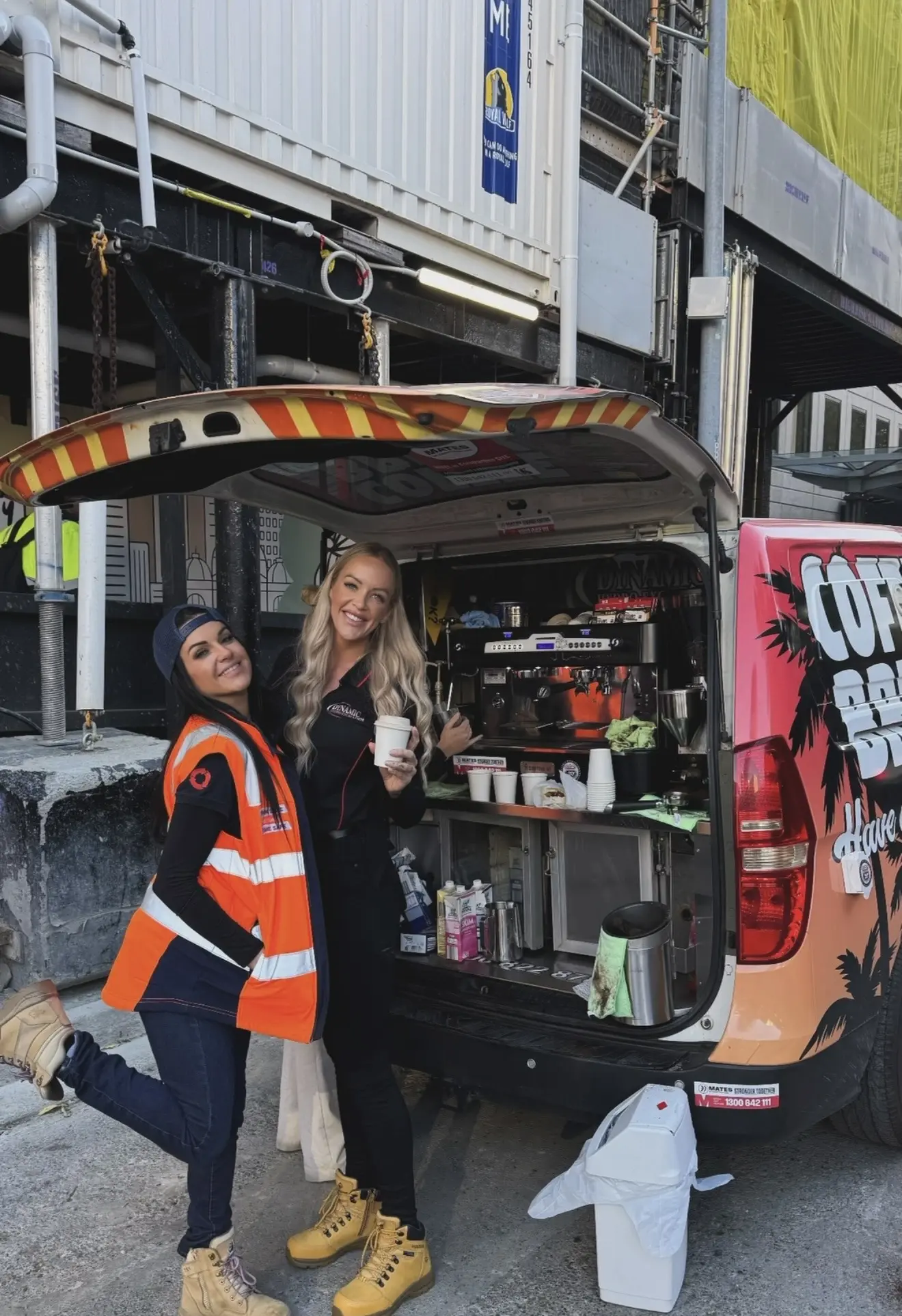 Two Dynamic team members smiling with takeaway coffees beside the open rear of the Coffee Break van, its espresso machine set up under scaffolding on a construction site