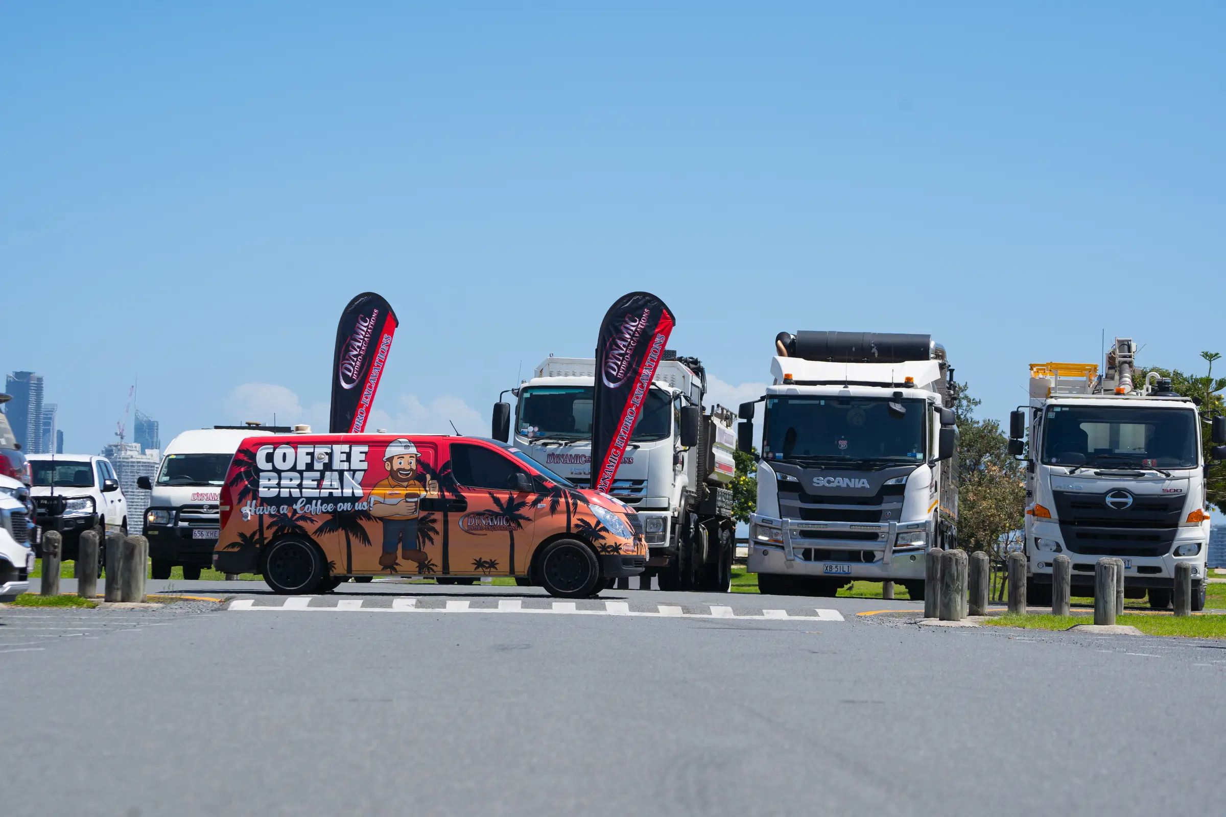 The Dynamic Coffee Break van parked in front of Dynamic vacuum excavation trucks and Dynamic flags, with a city skyline in the distance
