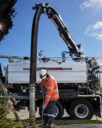 Dynamic operator in orange high-visibility clothing and a hard hat guiding a suction hose beside a Dynamic vacuum tanker with the boom extended overhead