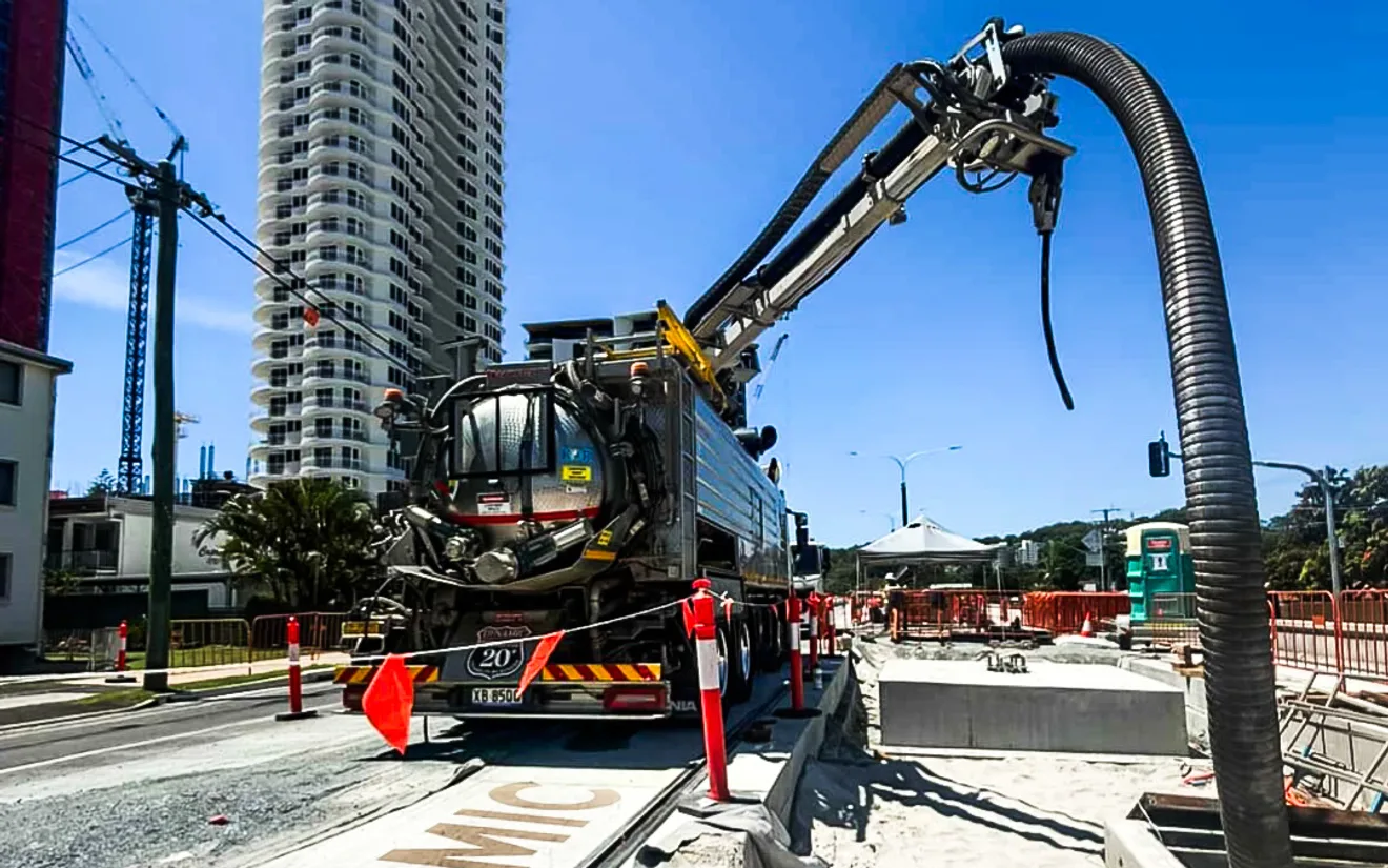 A Dynamic vacuum excavation truck with its boom and suction hose extended over an open roadside trench containing precast concrete stormwater structures