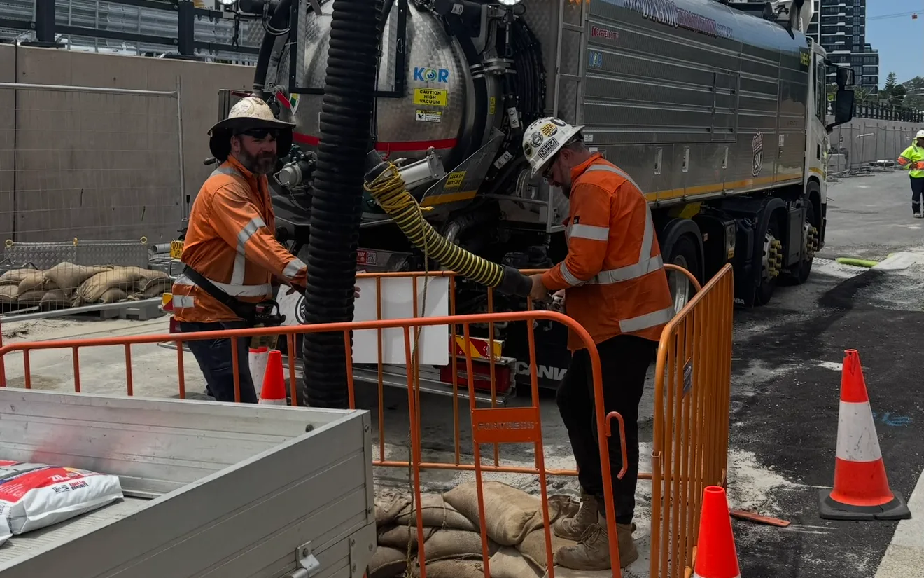 Two Dynamic crew in orange hi-vis and hard hats guiding a vacuum excavation suction hose beside a Dynamic vacuum truck to expose underground services in a barricaded city street